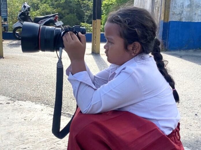 bocah perempuan berseragam sekolah dasar (SD) sedang memotret menggunakan kamera DSLR layaknya seorang fotografer.